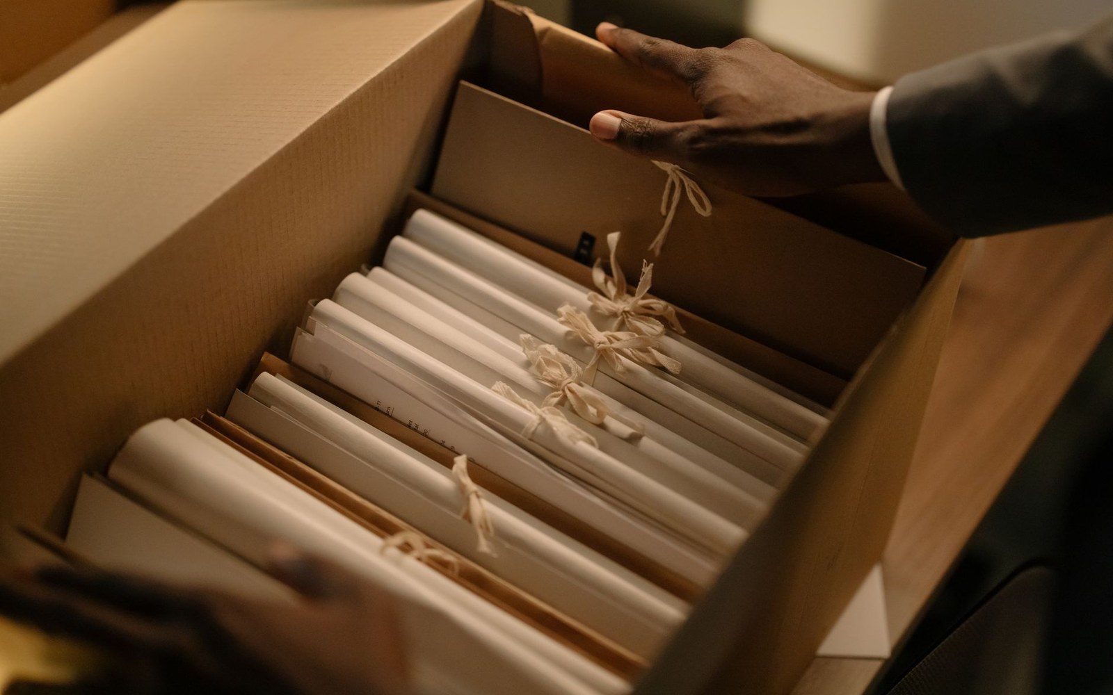 An open archive box containing printed campaign materials, an adult researcher's hands visible lifting one folder