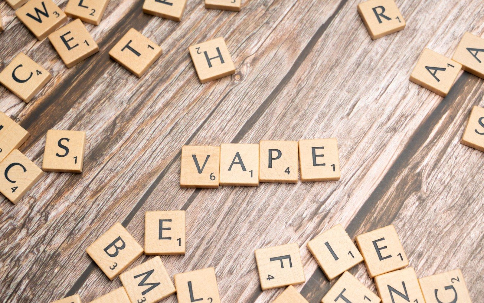 Scrabble tiles spelling "vape" surrounded by scattered letter tiles on a wooden surface